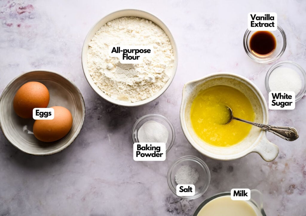 Bowls of baking ingredients for waffles&mdash;including eggs, all-purpose flour, baking powder, salt, melted butter, milk, white sugar, and vanilla extract&mdash;are neatly arranged on a countertop.