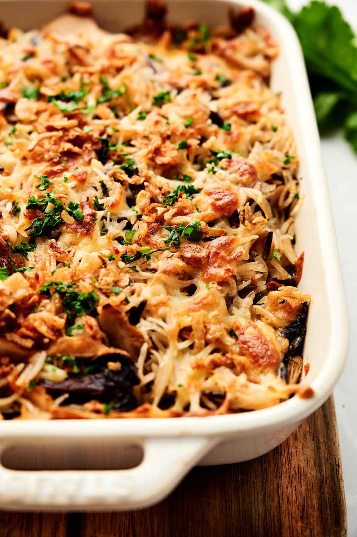 A Wild Rice Casserole topped with crispy fried onions and chopped parsley in a rectangular white baking dish on a wooden board.