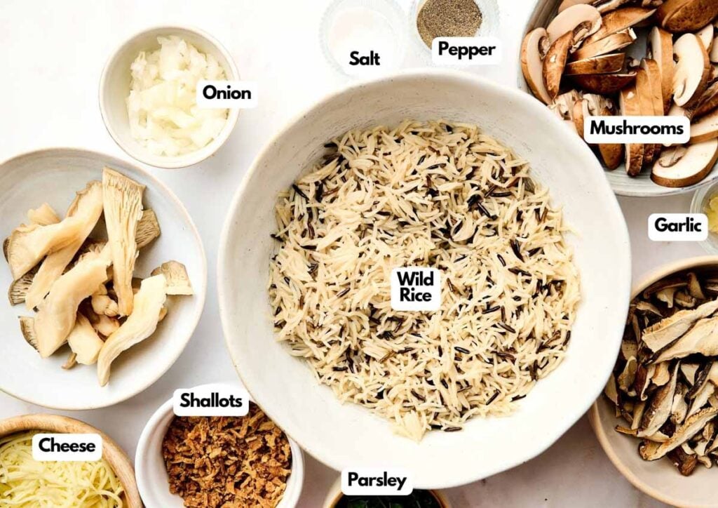 Bowls filled with labeled ingredients for Wild Rice Casserole, including wild rice, mushrooms, onion, garlic, shallots, cheese, parsley, salt, and pepper, arranged on a white surface.