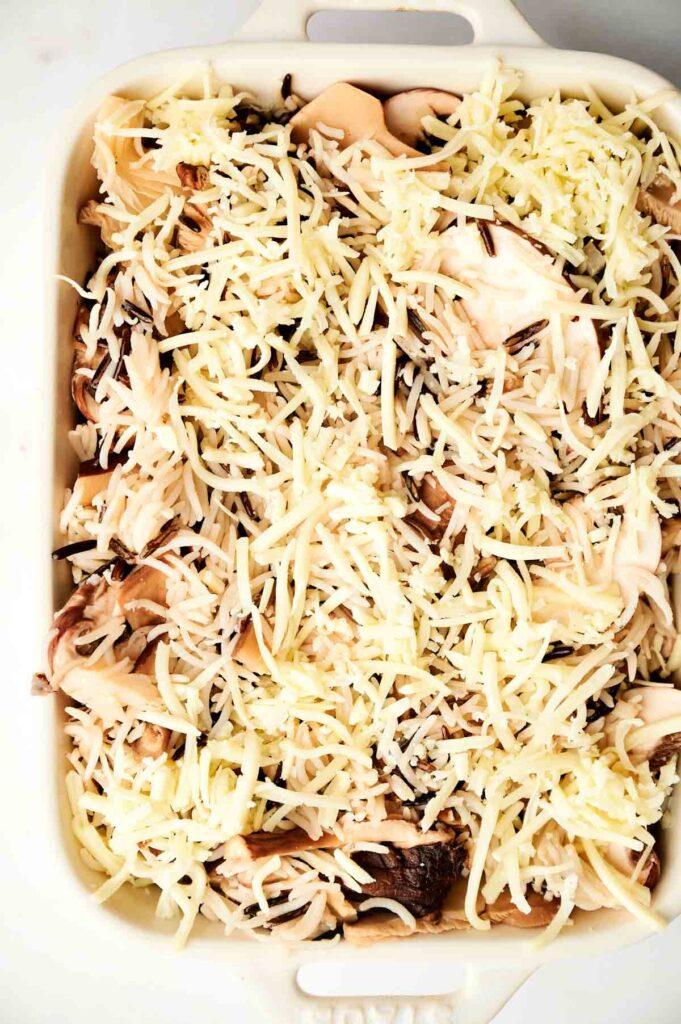 Wild Rice Casserole dish filled with shredded cheese, sliced mushrooms, and wild rice, ready to be baked.