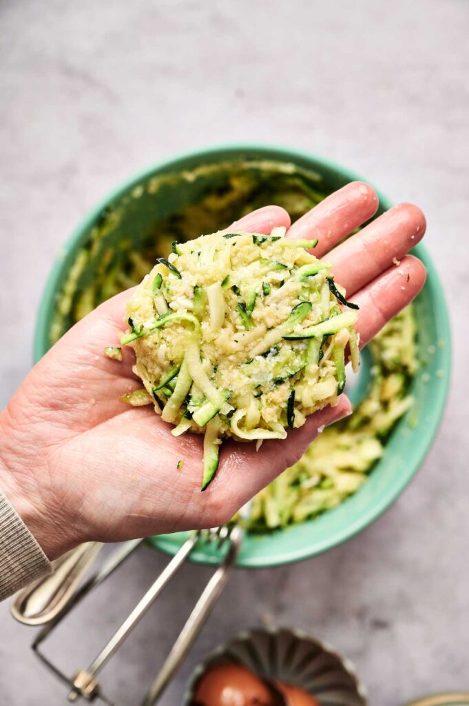 A hand holds a raw Air Fryer Zucchini Fritters patty over a green bowl filled with shredded zucchini mixture, with eggs and a whisk nearby.