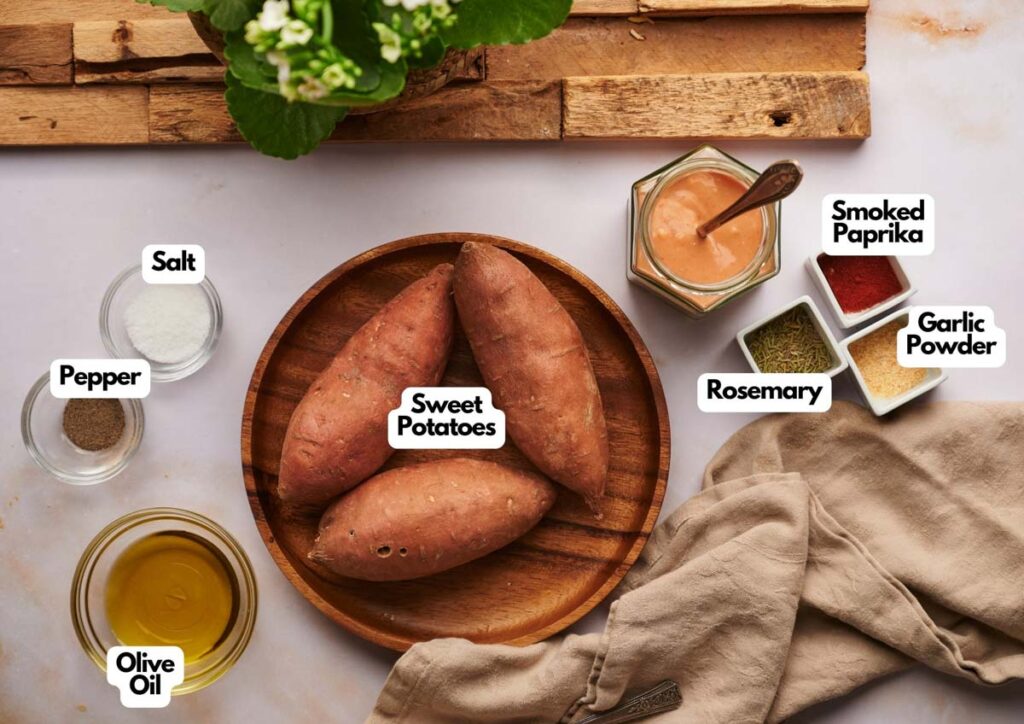 A wooden bowl with three sweet potatoes, perfect for making Sweet Potato Wedges, is surrounded by small bowls of olive oil, salt, pepper, smoked paprika, rosemary, and garlic powder on a light surface.