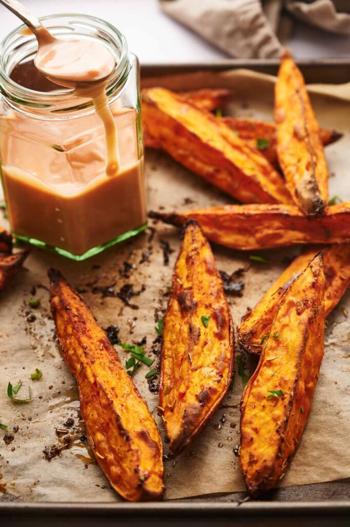 Baked sweet potato wedges on a parchment-lined tray, garnished with herbs, sit next to a jar of creamy dipping sauce&mdash;perfect for enjoying these delicious sweet potato wedges as a savory snack or side.
