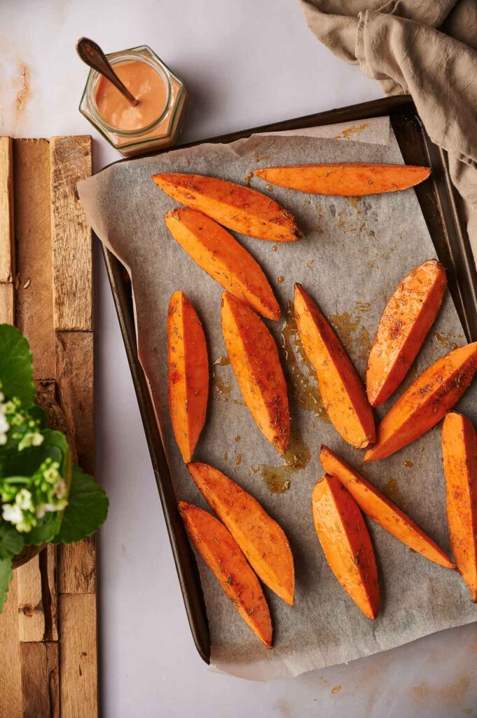 Baking tray with crispy Sweet Potato Wedges on parchment paper, served beside a jar of dipping sauce and a green plant.