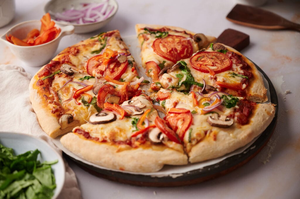A freshly baked Veggie Pizza with mushrooms, tomatoes, onions, and spinach sits on a round pizza pan, surrounded by bowls of fresh ingredients.