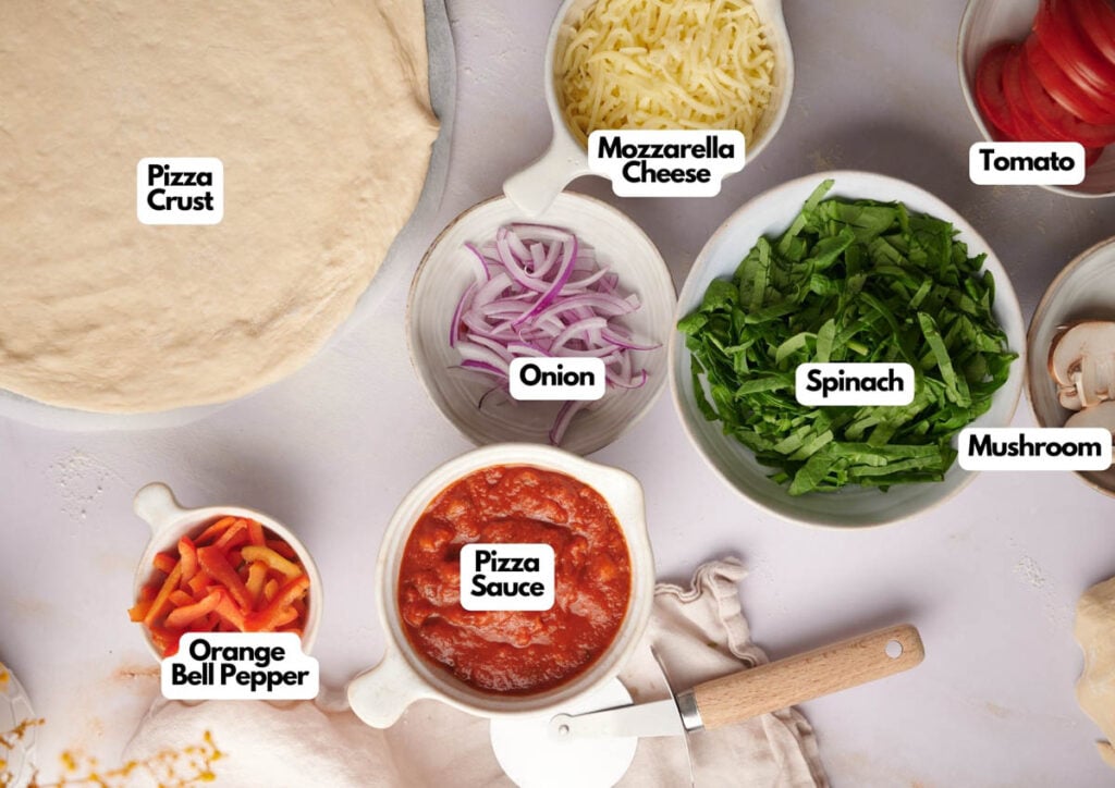 Veggie Pizza-making ingredients arranged on a counter: pizza crust, mozzarella cheese, tomato, mushroom, spinach, onion, orange bell pepper, and pizza sauce in labeled bowls.