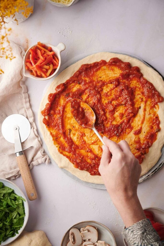 A hand spreads pizza sauce on pizza dough with a spoon; ingredients for a vegetarian pizza, including a pizza cutter, chopped vegetables, and toppings, are nearby on a light surface.