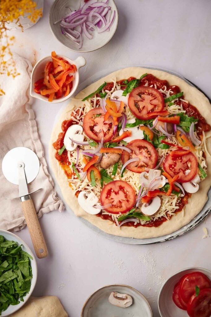 Unbaked vegetarian pizza topped with sliced tomatoes, mushrooms, onions, peppers, spinach, and cheese, surrounded by bowls of ingredients and a pizza cutter on a light surface.