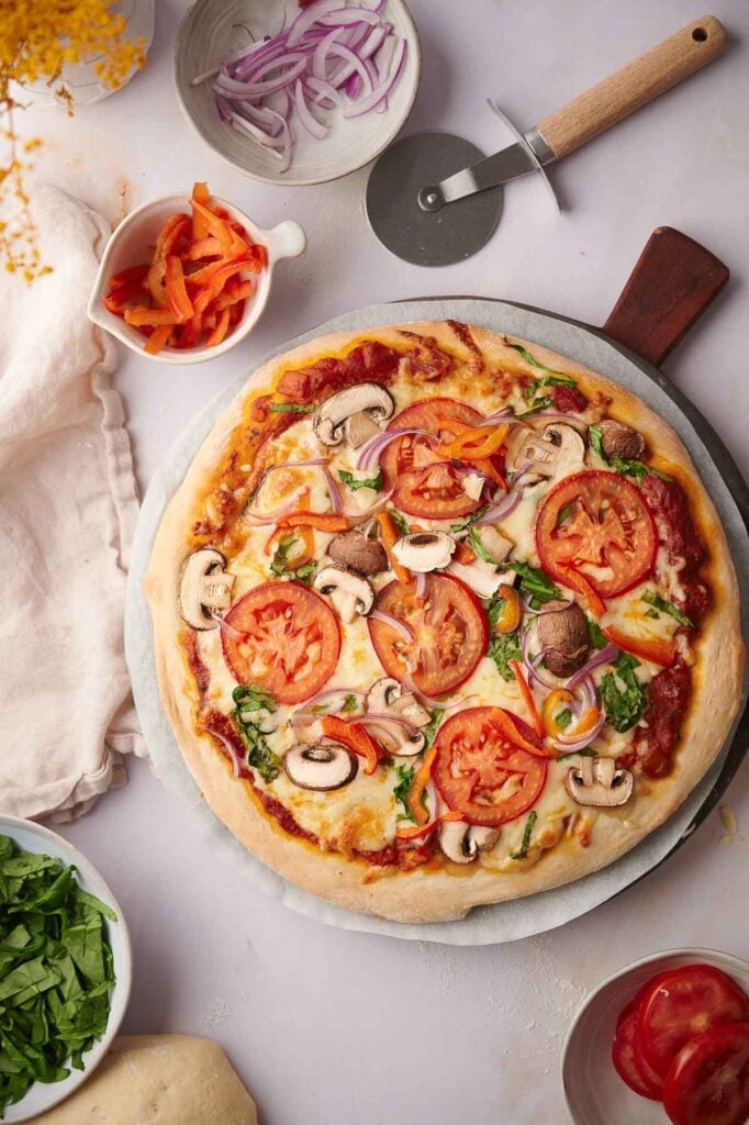 A freshly baked Veggie Pizza on a round tray, topped with tomatoes, mushrooms, spinach, onions, and peppers, surrounded by bowls of sliced vegetables and a pizza cutter.