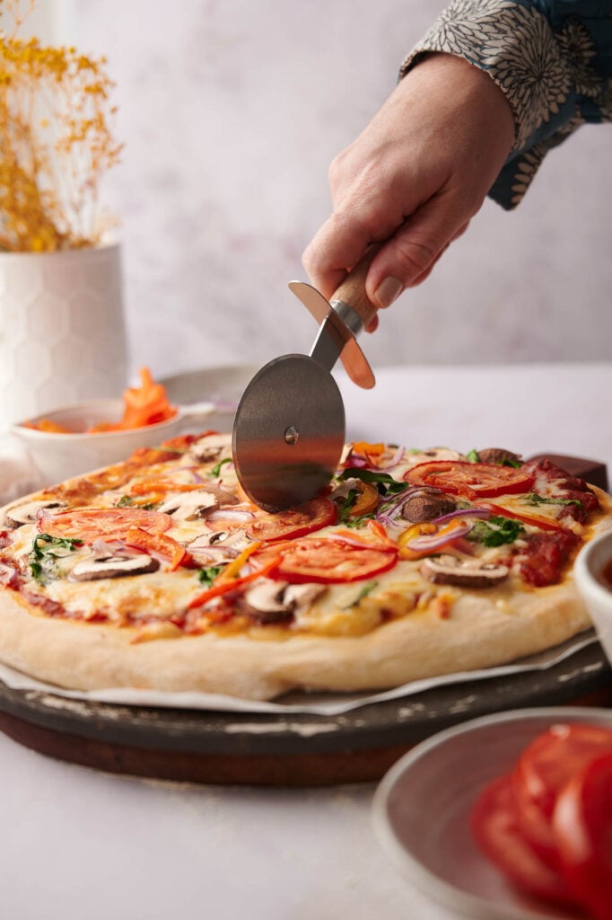 A hand uses a pizza cutter to slice a delicious vegetarian pizza on a wooden board. Plates and a bowl of tomato slices are nearby.