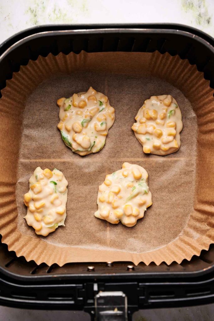 Four uncooked Air Fryer Corn Fritters rest on parchment paper inside an air fryer basket.