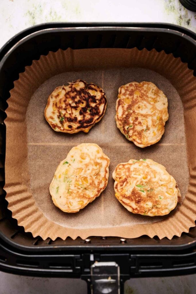 Four Air Fryer Corn Fritters rest on parchment paper inside an air fryer basket, partially browned and spaced apart for even cooking.