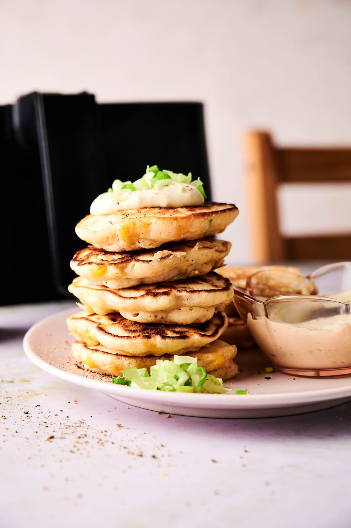 A stack of savory pancakes inspired by Air Fryer Corn Fritters, topped with a dollop of cream and chopped green onions, served on a plate with a side of dipping sauce.