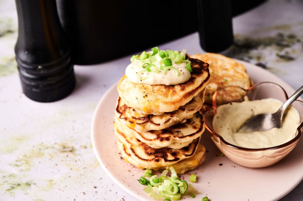 A stack of savory Air Fryer Corn Fritters topped with sour cream and chopped green onions, served on a plate next to a bowl of sauce with a spoon.
