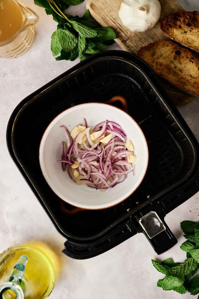 A bowl of sliced red onions and garlic sits inside an air fryer basket, ready to infuse flavor into your Air Fryer Pea and Mint Soup. Surrounding items include bread, fresh mint leaves, a glass pitcher, and a cup of broth—perfect complements for this vibrant dish.