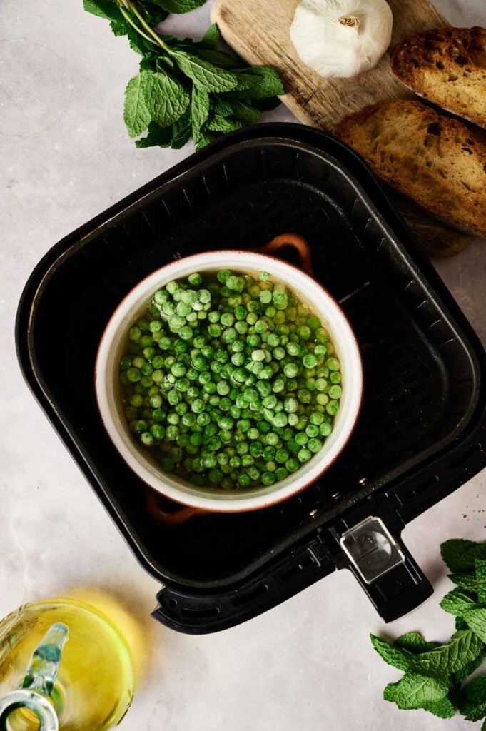 A bowl of frozen green peas sits in an air fryer basket, surrounded by fresh mint, garlic, toasted bread, and a jug of olive oil&mdash;perfect ingredients for a delicious Air Fryer Pea and Mint Soup.