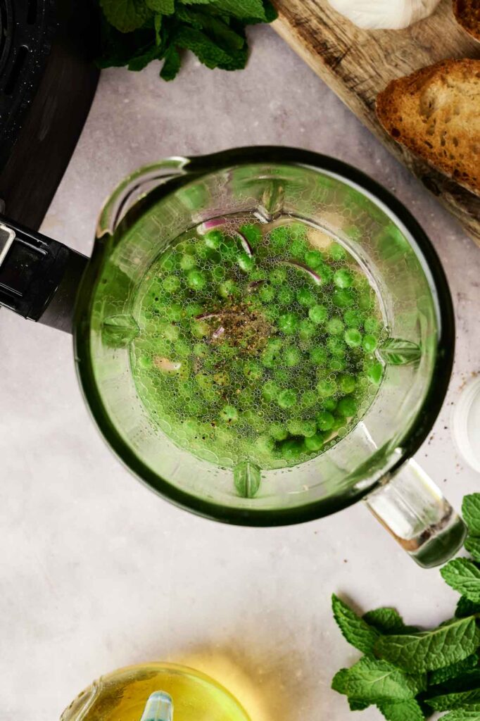 A top-down view of a blender with ingredients for Air Fryer Pea and Mint Soup—green peas, spices, and herbs—surrounded by toasted bread, mint leaves, and a jar of oil on a kitchen counter.