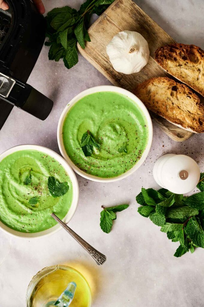 Two bowls of Air Fryer Pea and Mint Soup, garnished with fresh mint leaves, served alongside toasted bread, garlic cloves, more mint, and a pepper grinder on a light surface.