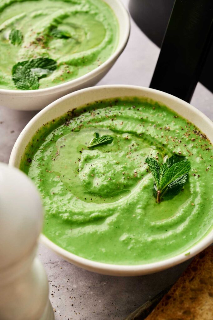 A bowl of Air Fryer Pea and Mint Soup garnished with mint leaves and black pepper, placed on a light-colored surface next to another similar bowl.