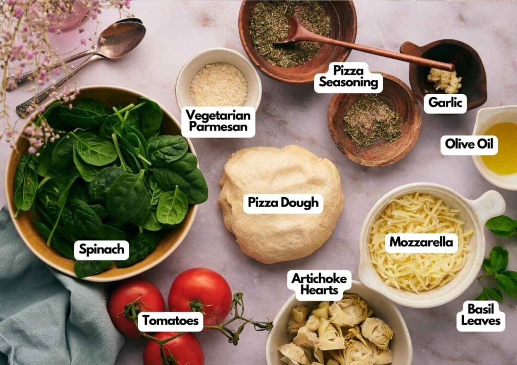 Labeled Artichoke Pizza ingredients on a table: spinach, tomatoes, artichoke hearts, pizza dough, mozzarella, vegetarian parmesan, pizza seasoning, garlic, olive oil, and basil leaves.