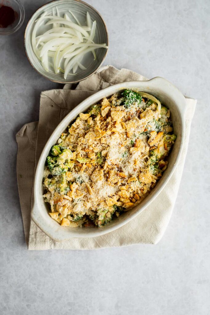 An oval casserole dish filled with a cheesy Broccoli Casserole, topped with golden breadcrumbs, sits on a beige cloth next to a bowl of sliced onions.
