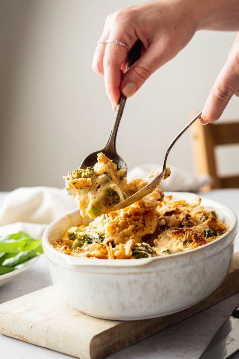 A hand uses two spoons to serve cheesy Broccoli Casserole and pasta from a white ceramic dish.