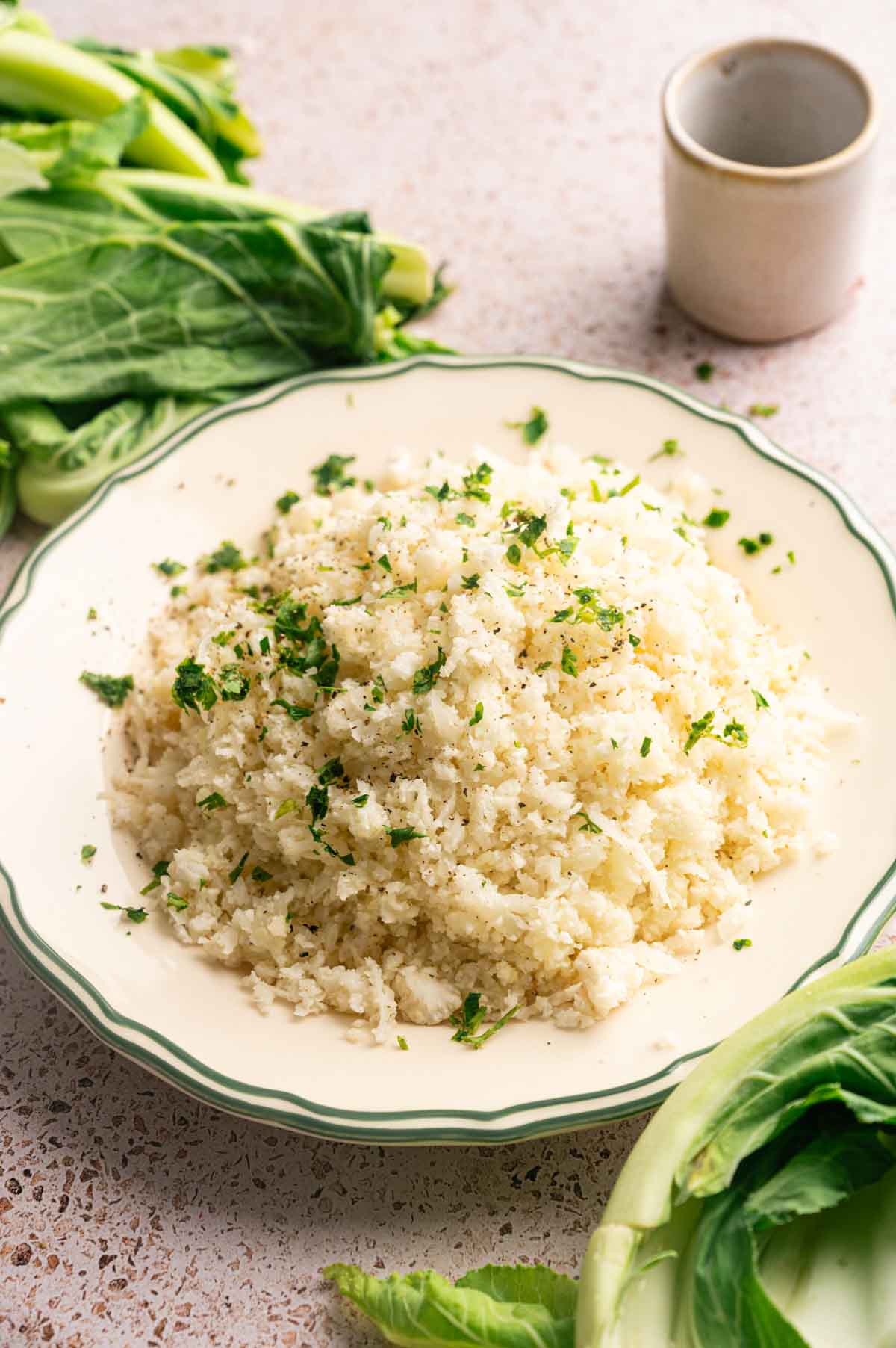 A plate of flavorful Cauliflower Rice garnished with chopped herbs, surrounded by fresh leafy greens on a speckled countertop.