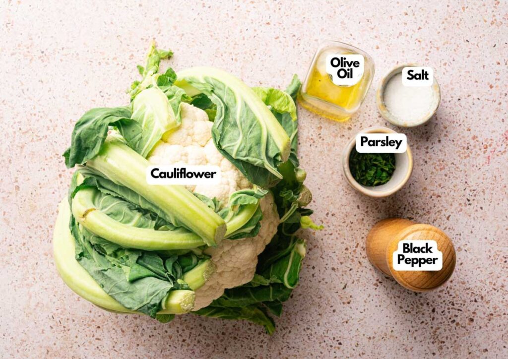 A head of cauliflower sits beside bowls of olive oil, salt, parsley, and a wooden pepper grinder labeled with their names&mdash;perfect ingredients for making homemade cauliflower rice.