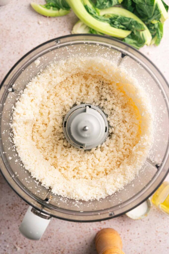 A food processor holds finely riced cauliflower, pulsed down to rice-sized pieces.