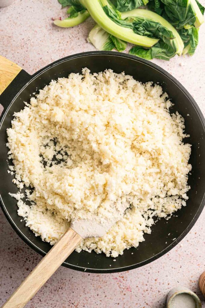 A skillet filled with cooked cauliflower rice sits on a countertop, a wooden spatula resting inside, with leafy greens in the background. This delicious cauliflower rice makes for a healthy and vibrant meal option.