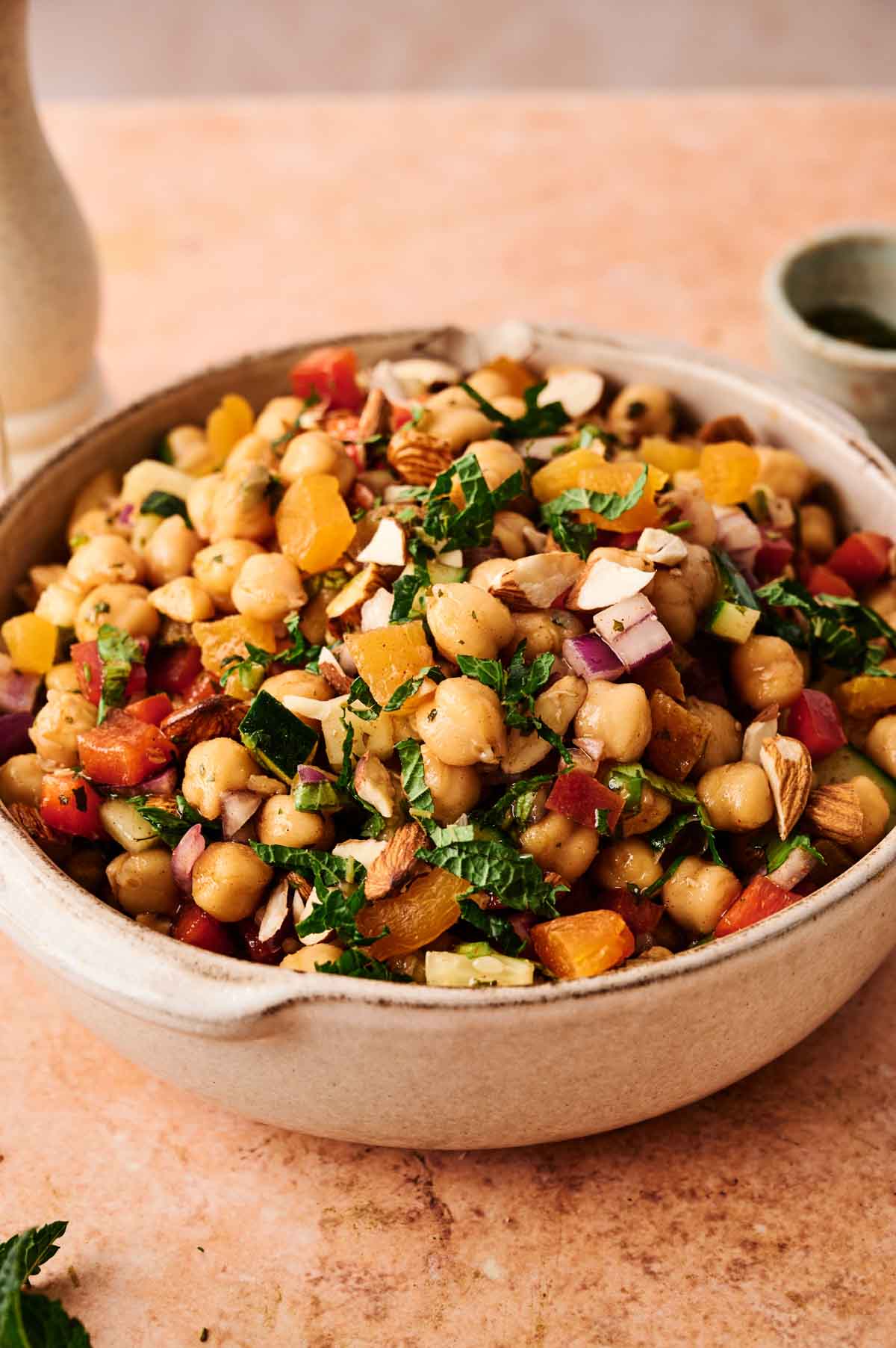 A vibrant Chickpea Salad featuring chopped greens, red onions, diced tomatoes, dried apricots, and sliced almonds sits on a light brown surface.