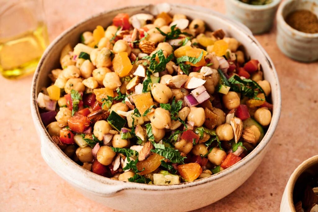 A bowl of Chickpea Salad with chopped vegetables, dried apricots, nuts, and fresh herbs sits on a light-colored surface.