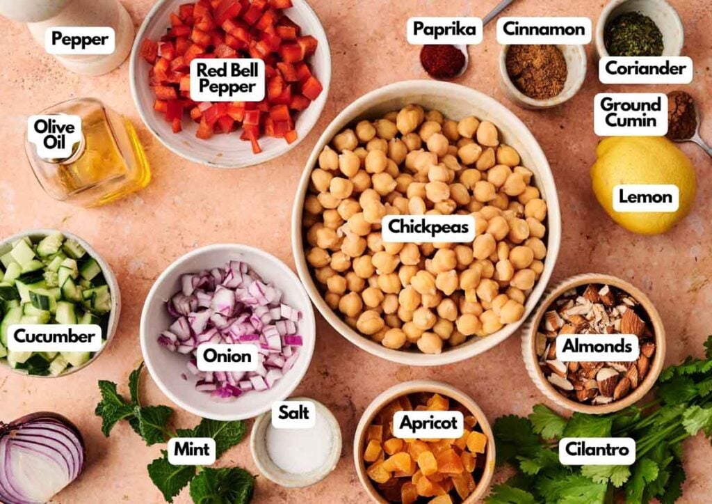 A variety of labeled Chickpea Salad ingredients on a countertop, including red bell pepper, cucumber, onion, almonds, apricot, spices, herbs, olive oil, and a lemon.