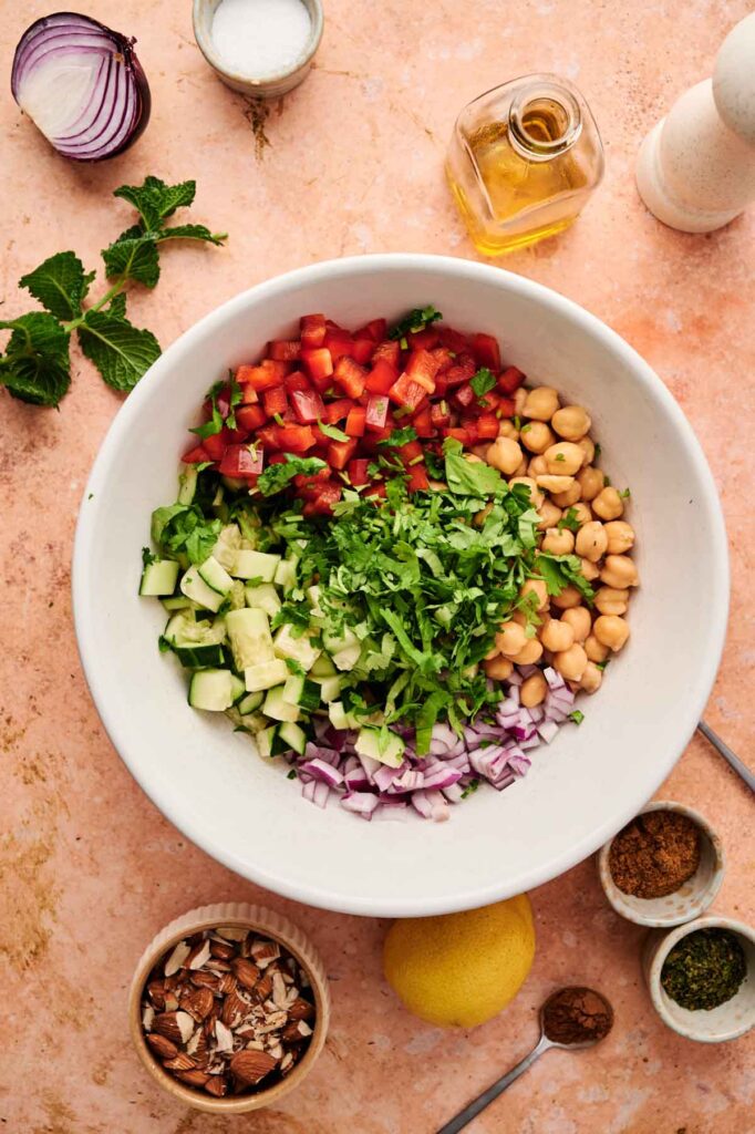 A bowl holds sections of diced vegetables and chickpeas, with almonds, lemon, and spices arranged around it.