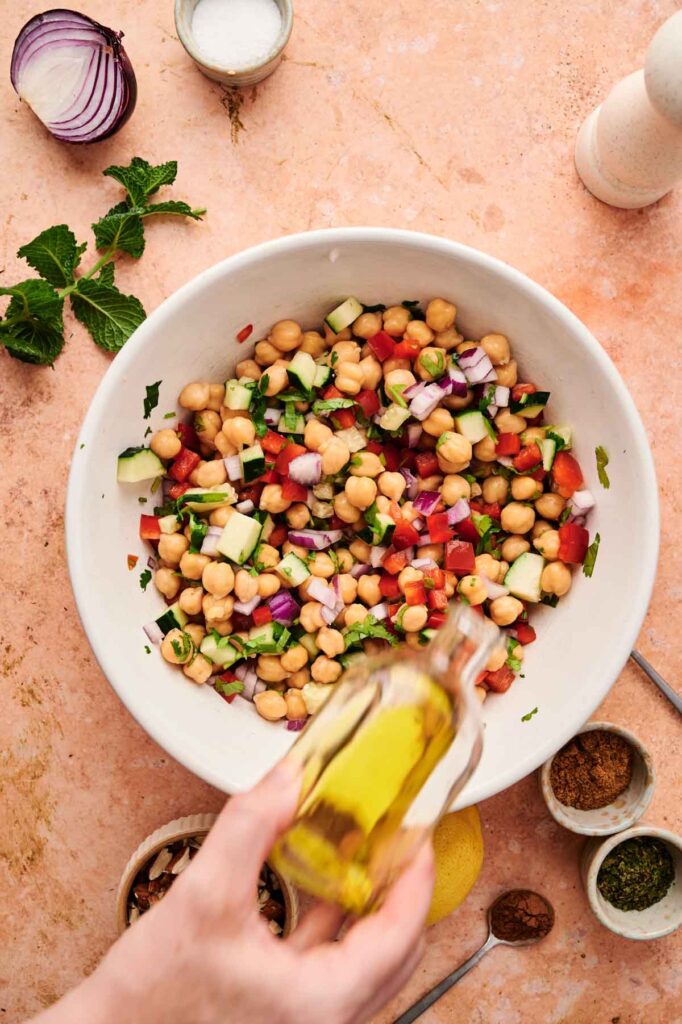 A hand drizzles olive oil into a bowl of tossed chickpea salad ingredients.