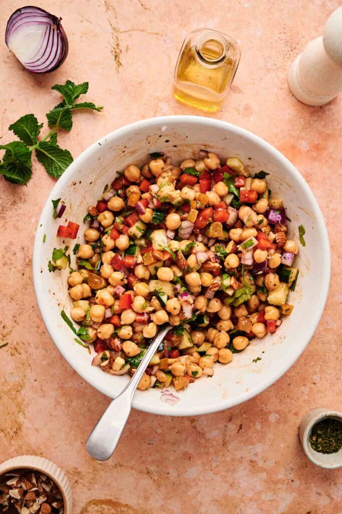 A bowl of Chickpea Salad with chopped vegetables and herbs sits on a countertop, surrounded by a bottle of oil, pepper grinder, mint leaves, and other fresh ingredients.