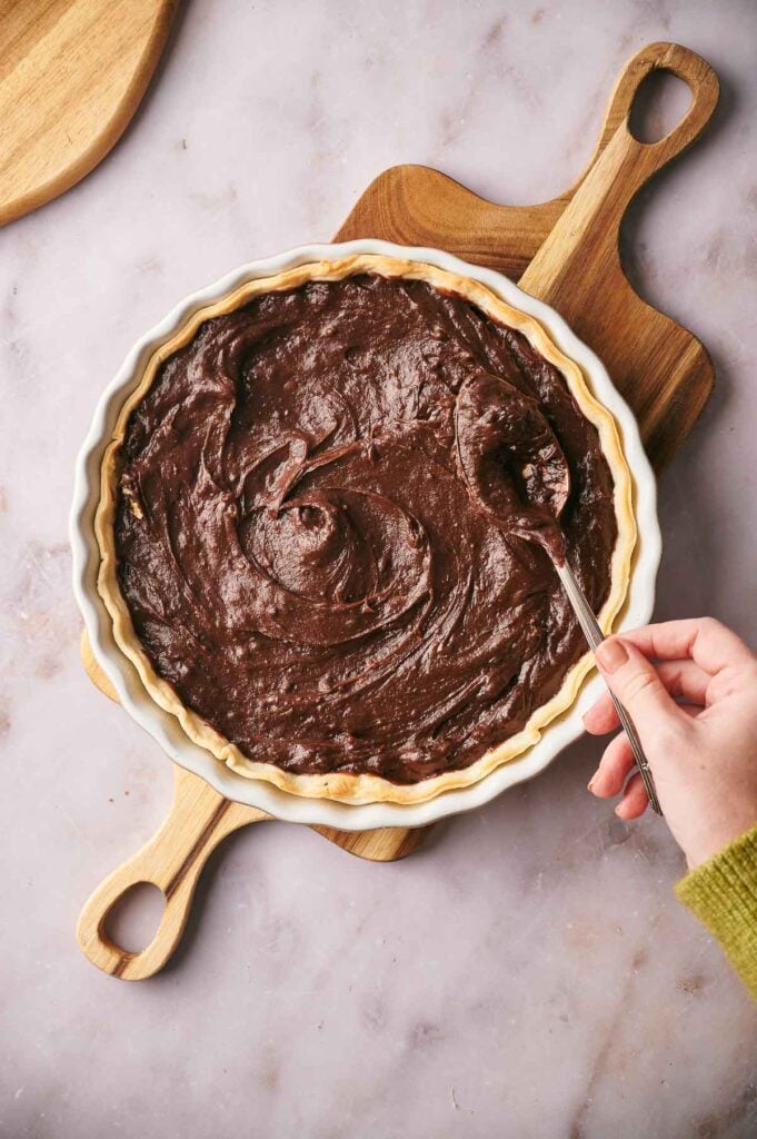 A hand spreads rich chocolate filling into a pie crust with a spoon, preparing a delicious Chocolate Pie on a wooden cutting board atop a marble surface.