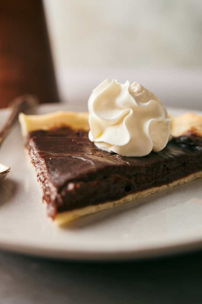 A slice of Chocolate Pie topped with a swirl of whipped cream sits on a plate next to a spoon, tempting any dessert lover.