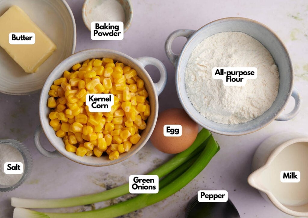 Bowls of kernel corn, all-purpose flour, and baking powder, plus butter, milk, egg, green onions, salt, and pepper arranged on a table&mdash;all labeled as the ingredients for delicious Air Fryer Corn Fritters.