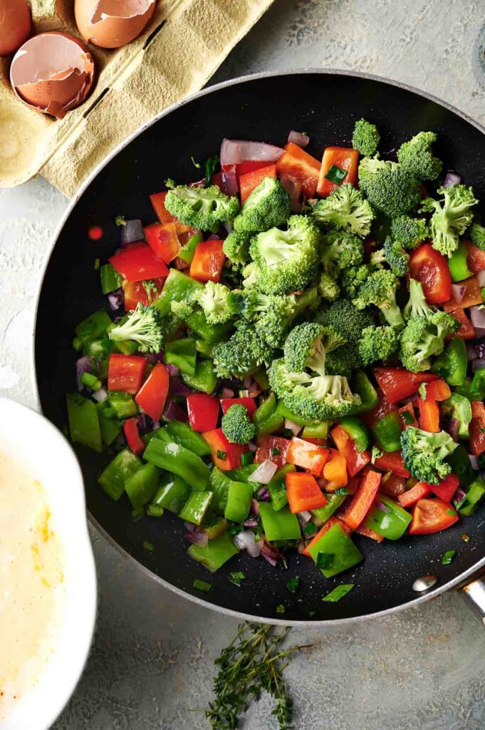 A frying pan filled with chopped broccoli, red bell peppers, green bell peppers, and onions sits on a countertop next to cracked eggshells—perfect ingredients for starting a flavorful egg casserole.