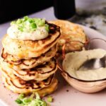 A stack of savory pancakes, reminiscent of Air Fryer Corn Fritters, topped with sour cream and chopped green onions sits beside a bowl of sauce with a spoon on a plate.