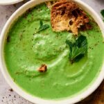 A bowl of creamy Air Fryer Pea and Mint Soup, garnished with mint leaves and cracked black pepper, served with pieces of toasted bread.