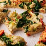 Close-up of an Artichoke Pizza topped with spinach and tomatoes. One slice is missing a bite, and the pizza rests on white parchment paper.