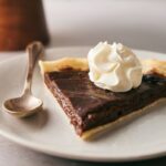 A slice of Chocolate Pie topped with whipped cream sits on a plate next to a spoon.