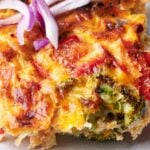 A slice of vegetable quiche, similar to an egg casserole, with visible pieces of broccoli, red bell pepper, and melted cheese, garnished with sliced red onion on top.