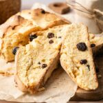 Sliced loaf of Irish Soda Bread with raisins on parchment paper, set on a wooden board with a small round jar and twine in the background.