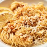 A plate of Lemon Pasta topped with grated cheese, black pepper, and red pepper flakes, garnished with two lemon slices.