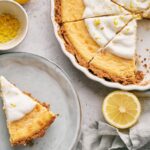 A Lemon Pie with a graham cracker crust and whipped topping is partially sliced in a dish; one slice is served on a plate beside a lemon half and a bowl of fresh lemon zest.
