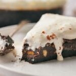 A slice of Mississippi Mud Pie brownie with nuts is topped with melting vanilla ice cream on a white plate, with a fork holding a bite nearby.