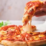 A hand lifts a cheesy slice from a freshly baked Neapolitan pizza topped with tomato sauce and mozzarella cheese.